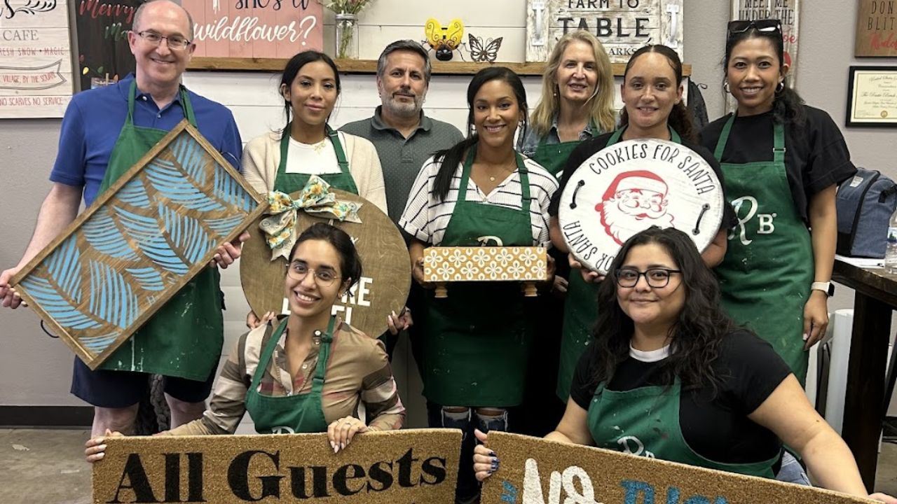 Group of adults posing at The Rustic Brush, holding handmade wooden signs and painted doormats 