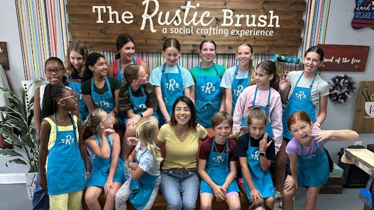 Group of smiling kids and a woman wearing aprons at The Rustic Brush crafting studio.