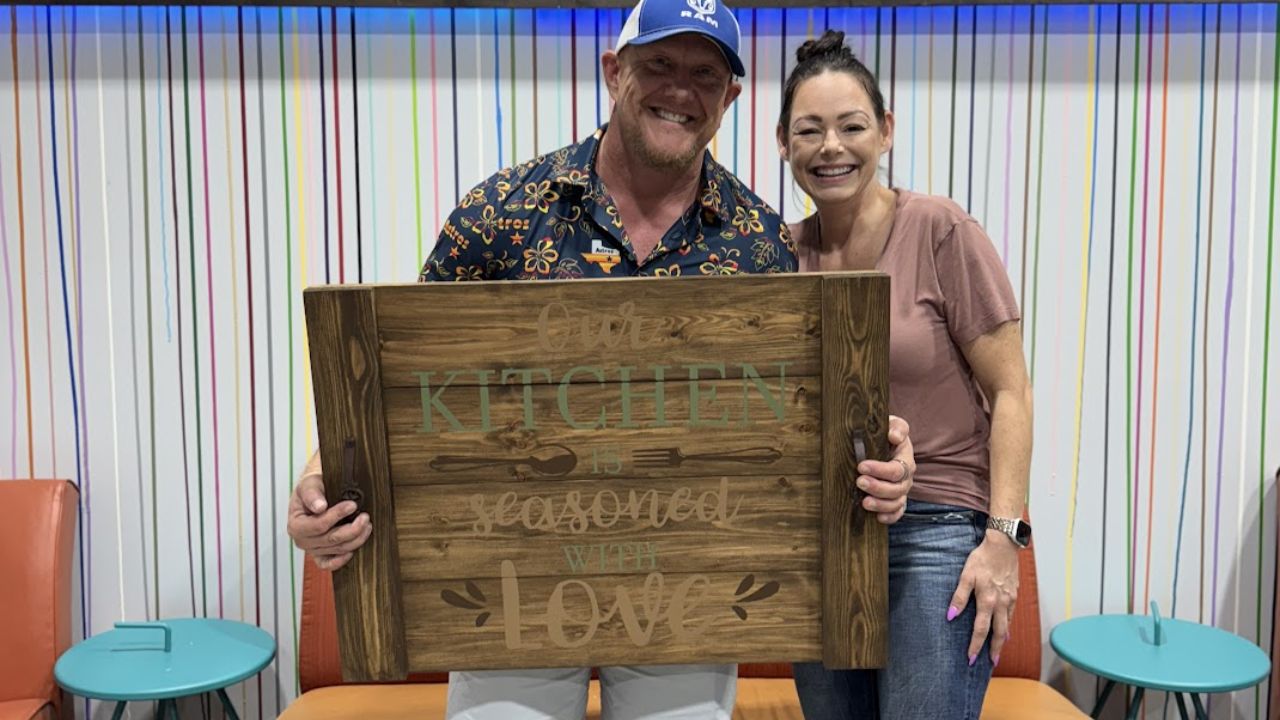 A couple smiling and holding a finished wooden home decor sign inside The Rustic Brush craft studio.