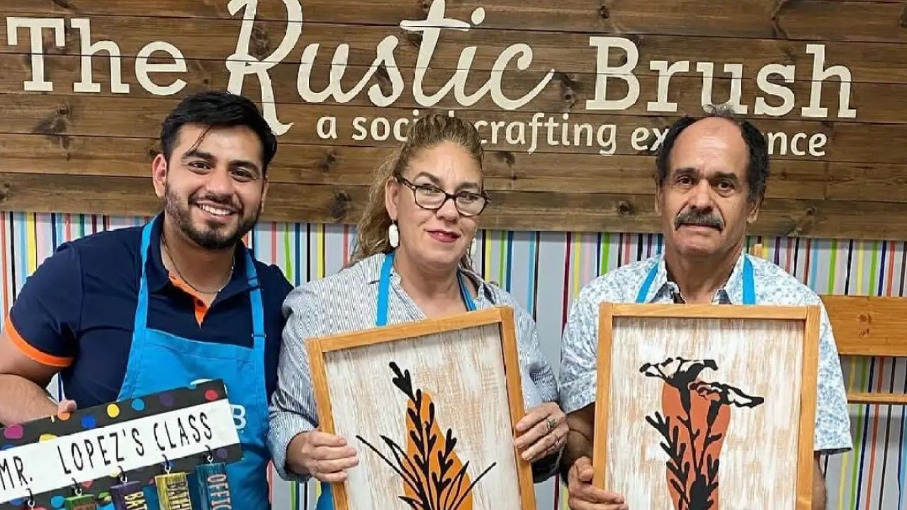 Three people wearing aprons holding handmade wooden wall art they created at The Rustic Brush DIY workshop.