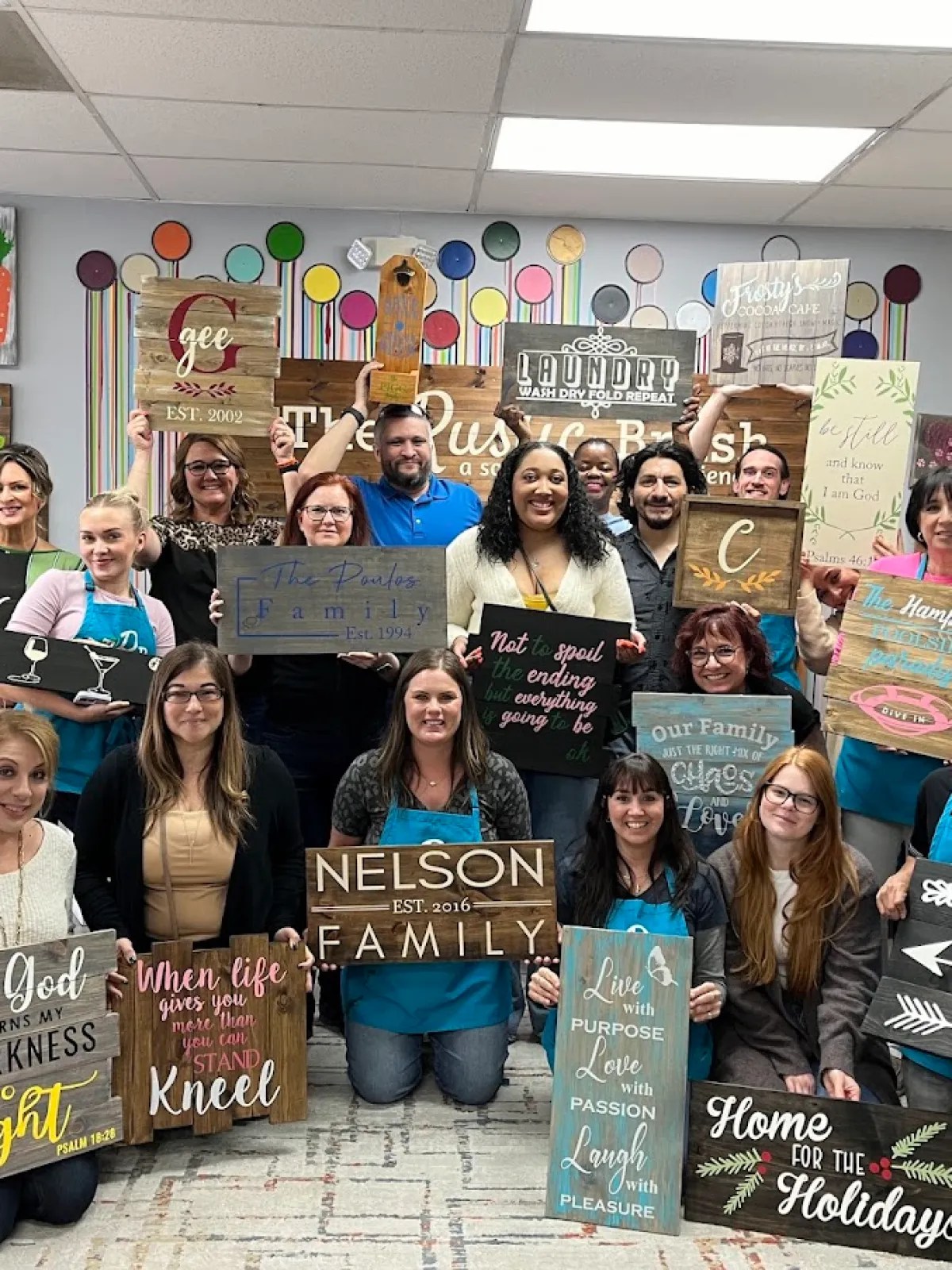 Large group of people posing with their personalized wooden signs after a DIY crafting workshop at The Rustic Brush.