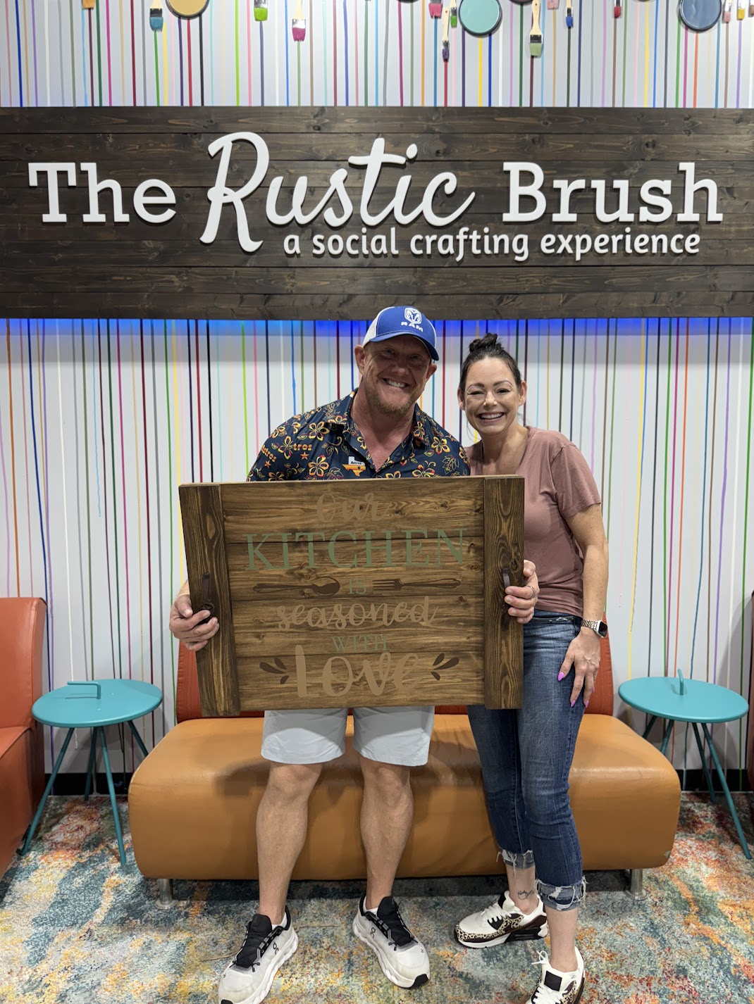 Couple smiling and holding a handmade wooden kitchen sign at The Rustic Brush crafting studio.