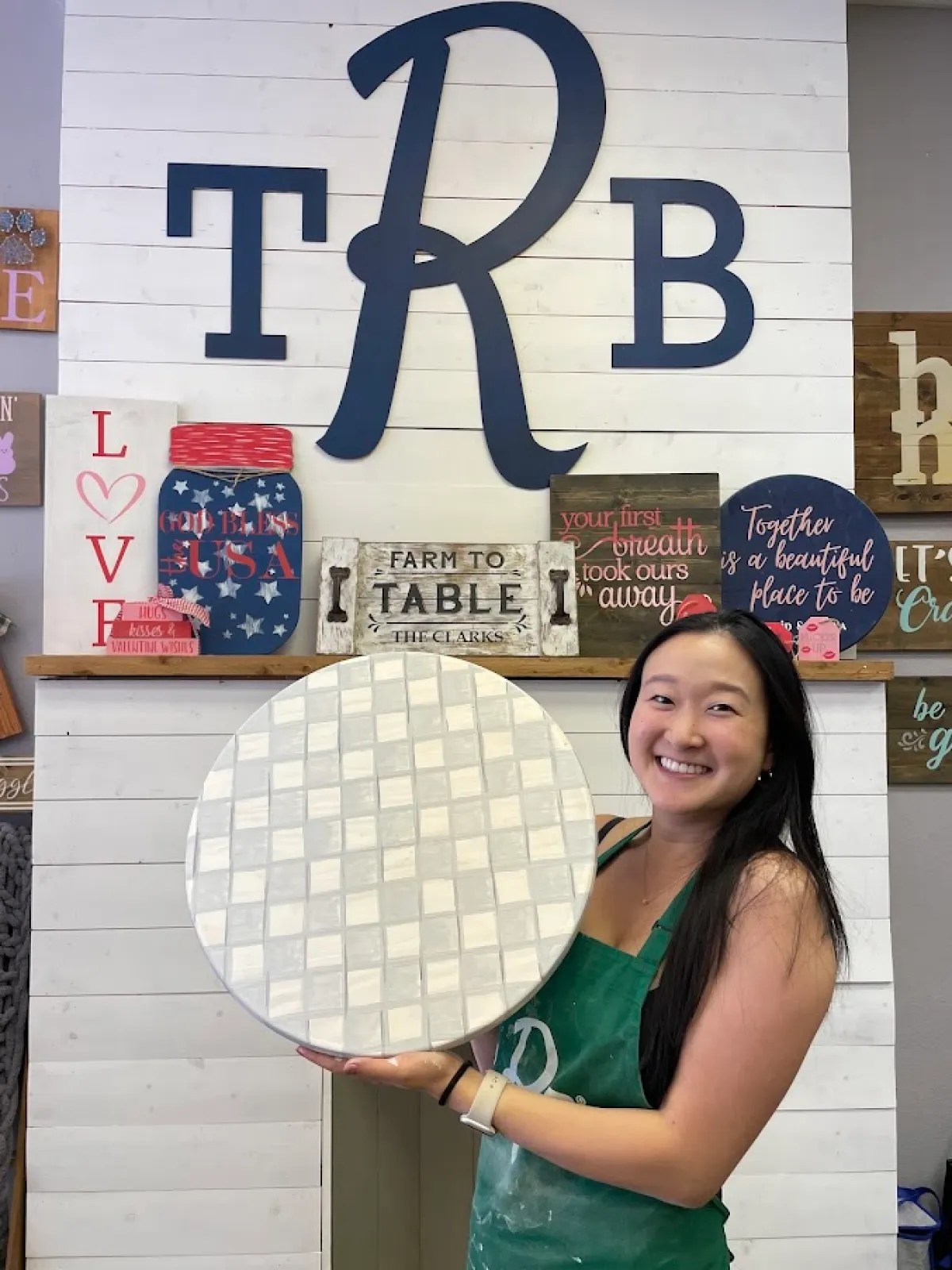 Smiling woman holding a round mosaic tile in front of a shiplap wall with decorative signs.