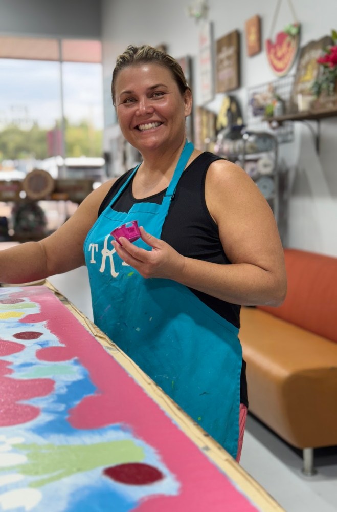 Woman painting a colorful wooden board while smiling inside a creative workshop.