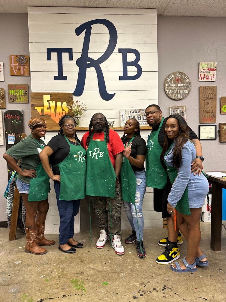 Six people in aprons posing in front of wall art with 'TRB' letters.