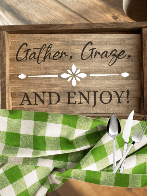 Wooden sign with 'Gather, Graze, and Enjoy!' above a green checkered cloth and utensils.