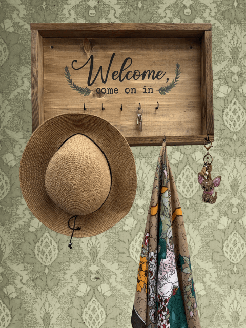 Wooden sign with 'Welcome' above hooks holding a hat and scarf on patterned wallpaper.