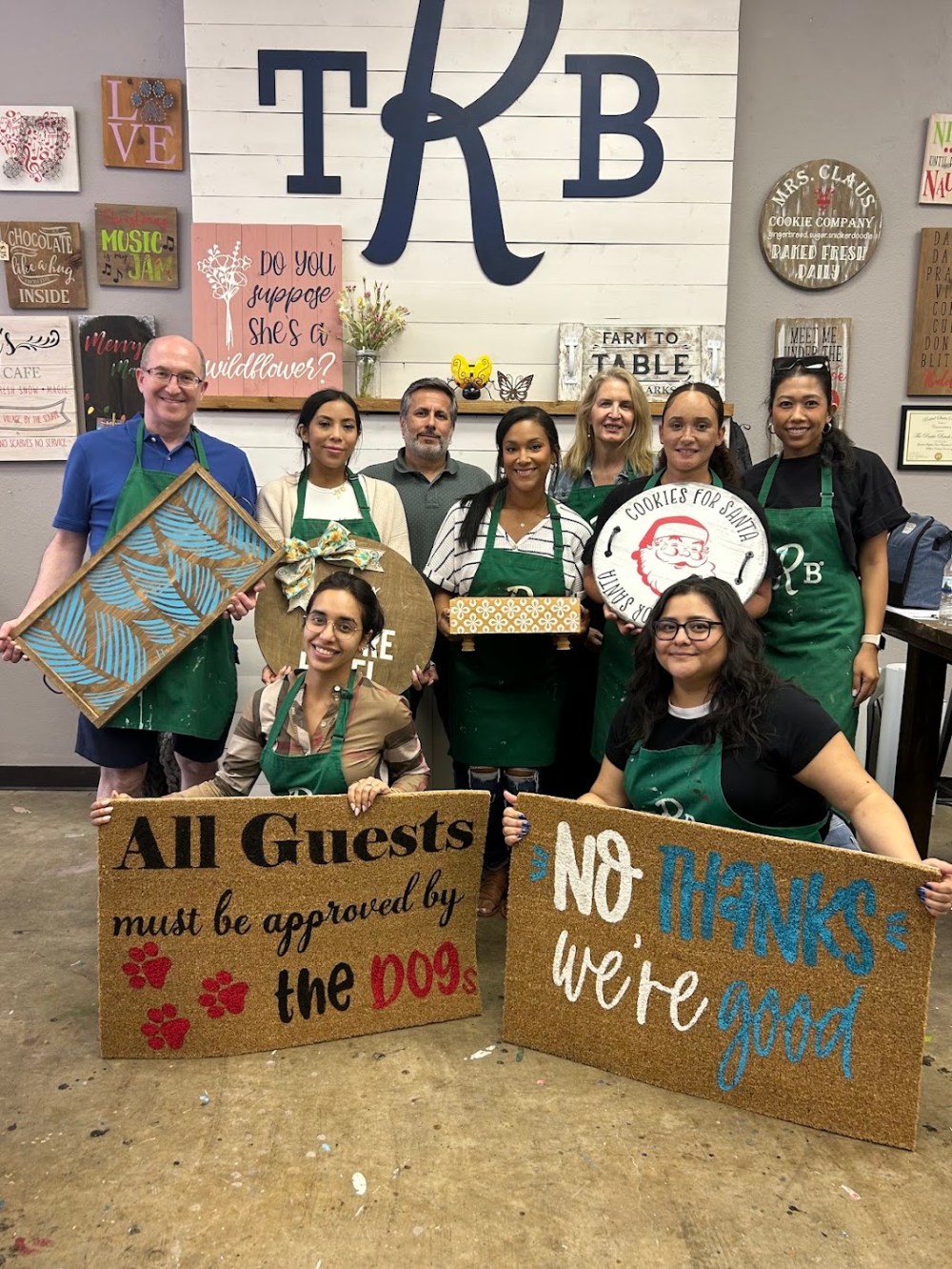 Five friends wearing green aprons smiling together before a fun DIY painting and woodworking workshop at The Rustic Brush craft studio.
