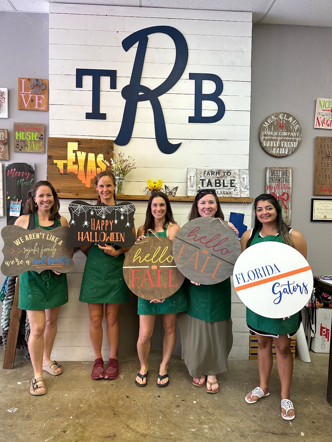 Five women displaying their hand-painted wooden signs in a craft studio.